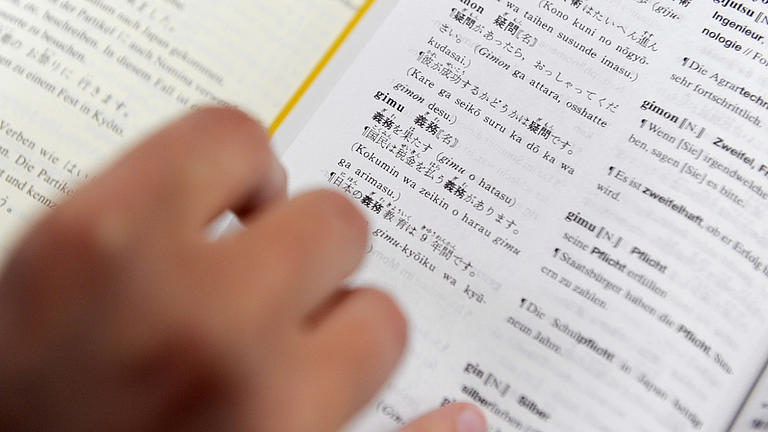 Nachaufnahme eines geöffneten japanisch-deutschen Wörterbuches, auf dessen Seite eine Hand ein Lesezeichen hält.