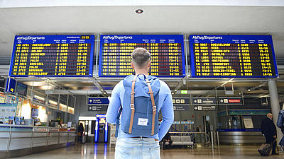 Aufnahme eines jungen Mannes von hinten, er hat kurze Haare, ist bekleidet mit einer blauen Jeans, einem blauen Langarmshirt, hat einen blauen Rucksack auf dem Rücken und steht vor den Anzeigetafeln einer Abflughalle in einem Flughafen.