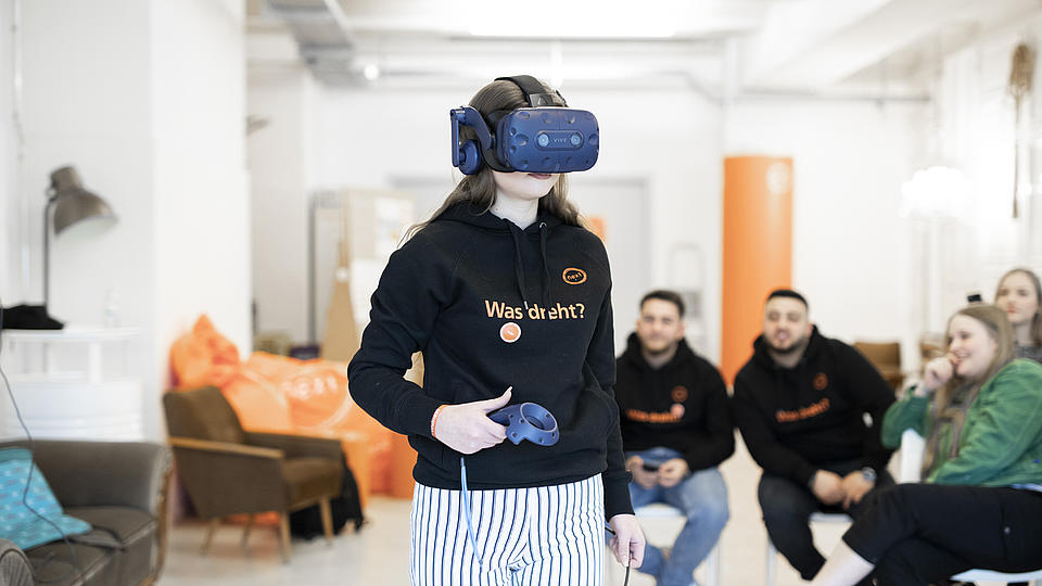 Eine Auszubildende entdeckt mit einer VR-Brille virtuelle Welten. Hinter ihr sitzen weitere Auszubildende in einem Stuhlkreis und beobachten sie.