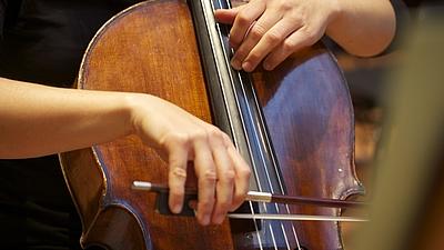 Nahaufnahme eines Cellos, das von einem Mann gehalten wird. Eine Hand hÃ¤lt einen Bogen Ã¼ber das Instrument. Die andere hält die Saiten fest.