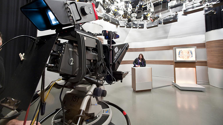  Nahaufnahme einer Fernsehkamera, die auf eine im Hintergrund stehende Frau gerichtet ist.