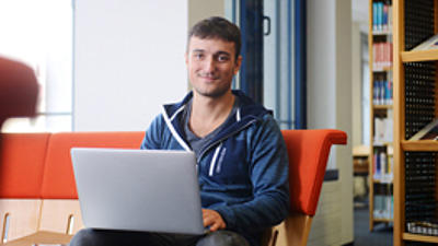 Ein junger Mann sitzt mit Laptop auf einer Couch.
