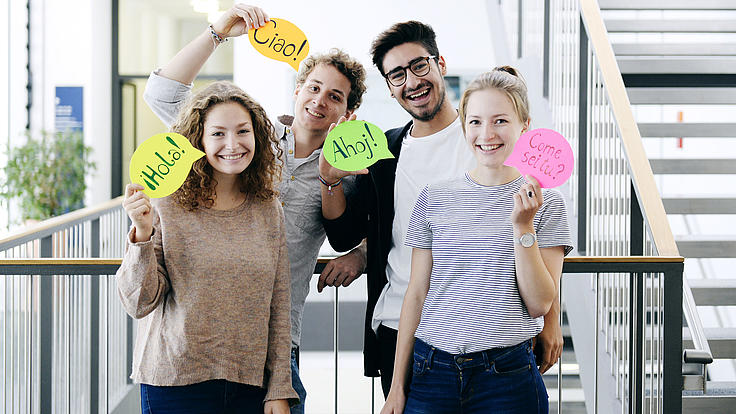 Vier junge Menschen zeigen Sprechblasen.Schilder. Auf diesen ist in vier Fremdsprachen das Wort Hallo zu lesen.