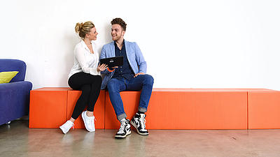 Eine junge Frau und ein junger Mann sitzen zusammen auf einem orangefarbenen Kasten neben einer blauen Couch.