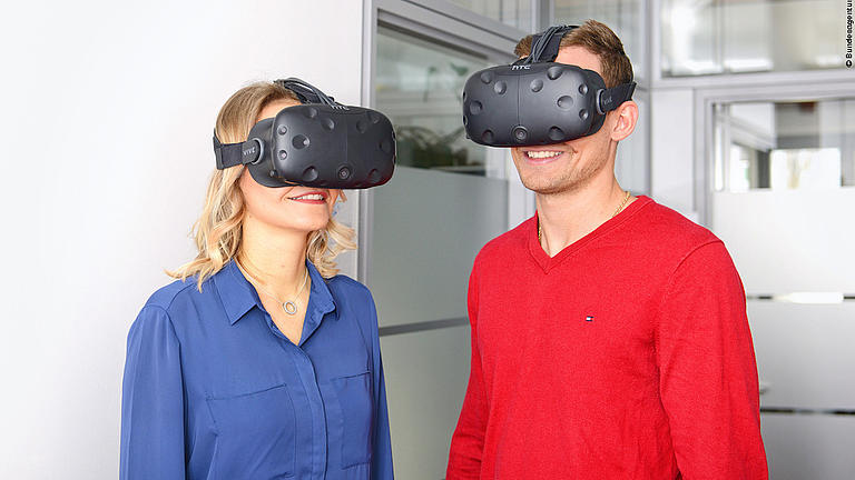 Ein Mann und eine Frau mit aufgesetzten Virtual-Reality-Brillen stehen in einem Raum und lächeln