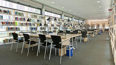 Blick in einen Lesesaal mit mehreren Tischreihen, an denen schwarze Stühle stehen.