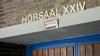 Nahaufnahme einer Raumbezeichnung in einem Hochschulgebäude. Über einer Holztür mit blauem Rahmen steht in Großbuchstaben