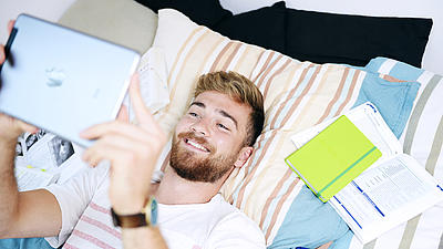 Student liegt mit Tablet und Büchern auf einem Bett