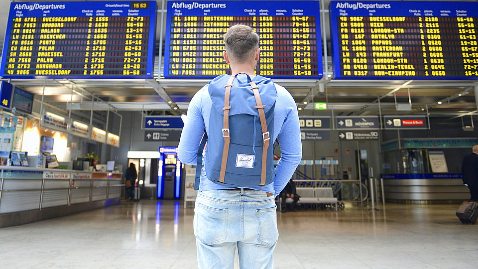 Ein junger Mann steht vor einer Abflugtafel am Flughafen.