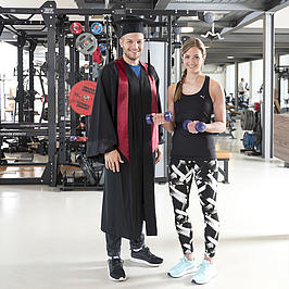 Zwei junge Menschen stehen in einem Fitnessstudio. Die junge Frau trägt Sportklamotten, der junge Mann einen Doktorhut und Robe.