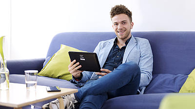 Ein junger Mann sitzt auf einer blauen Couch und hält ein Tablet in seinen Händen. Er hat ein Bein auf das andere gelegt und blickt lächelnd in die Kamera. 