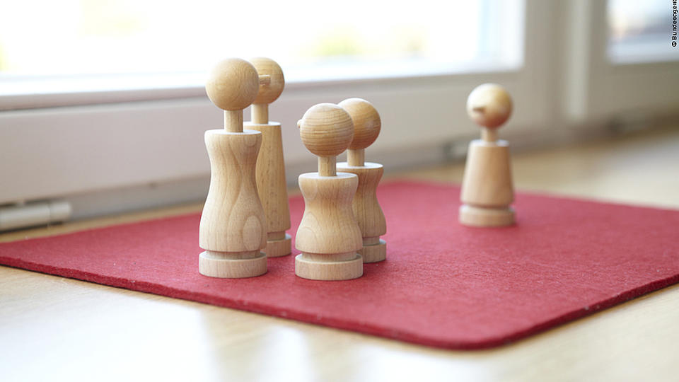 Auf einer quadratischen roten Filzunterlage mit abgerundeten Ecken stehen vier hölzerne Spielfiguren. Zwei große und zwei kleine Figuren stehen zusammen, etwas weiter hinten eine weitere kleine Holzfigur.